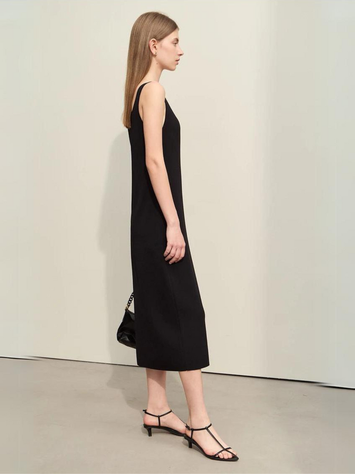 Profile view of a woman wearing a plain, minimalist, sleeveless midi dresses with a v-cut out in the neckline in black. She has on black strappy heels and a black bag to complete the look.