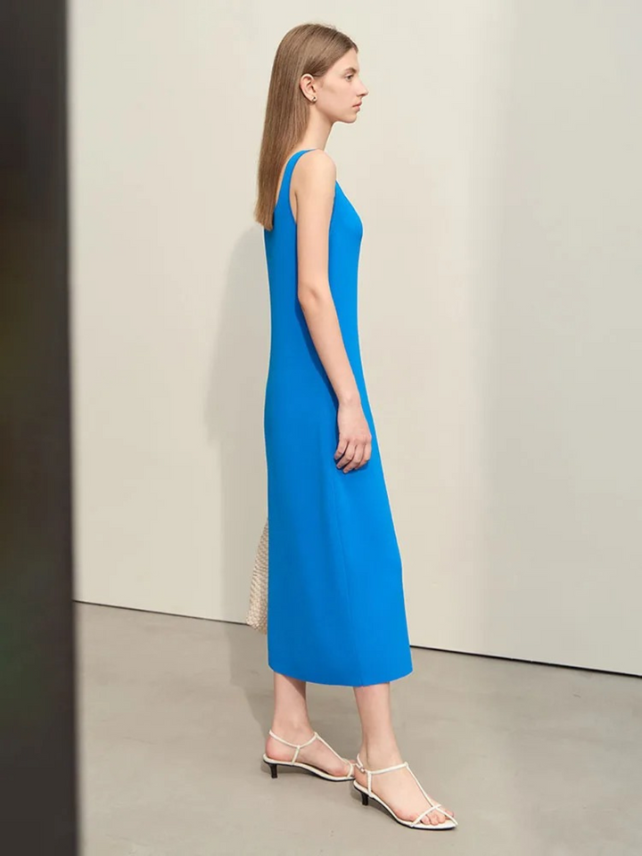 Profile view of A woman wearing a plain, minimalist, sleeveless midi dresses with a v-cut out in the neckline in blue. She has on a pair of white strappy heels and a white bag to complete the look.