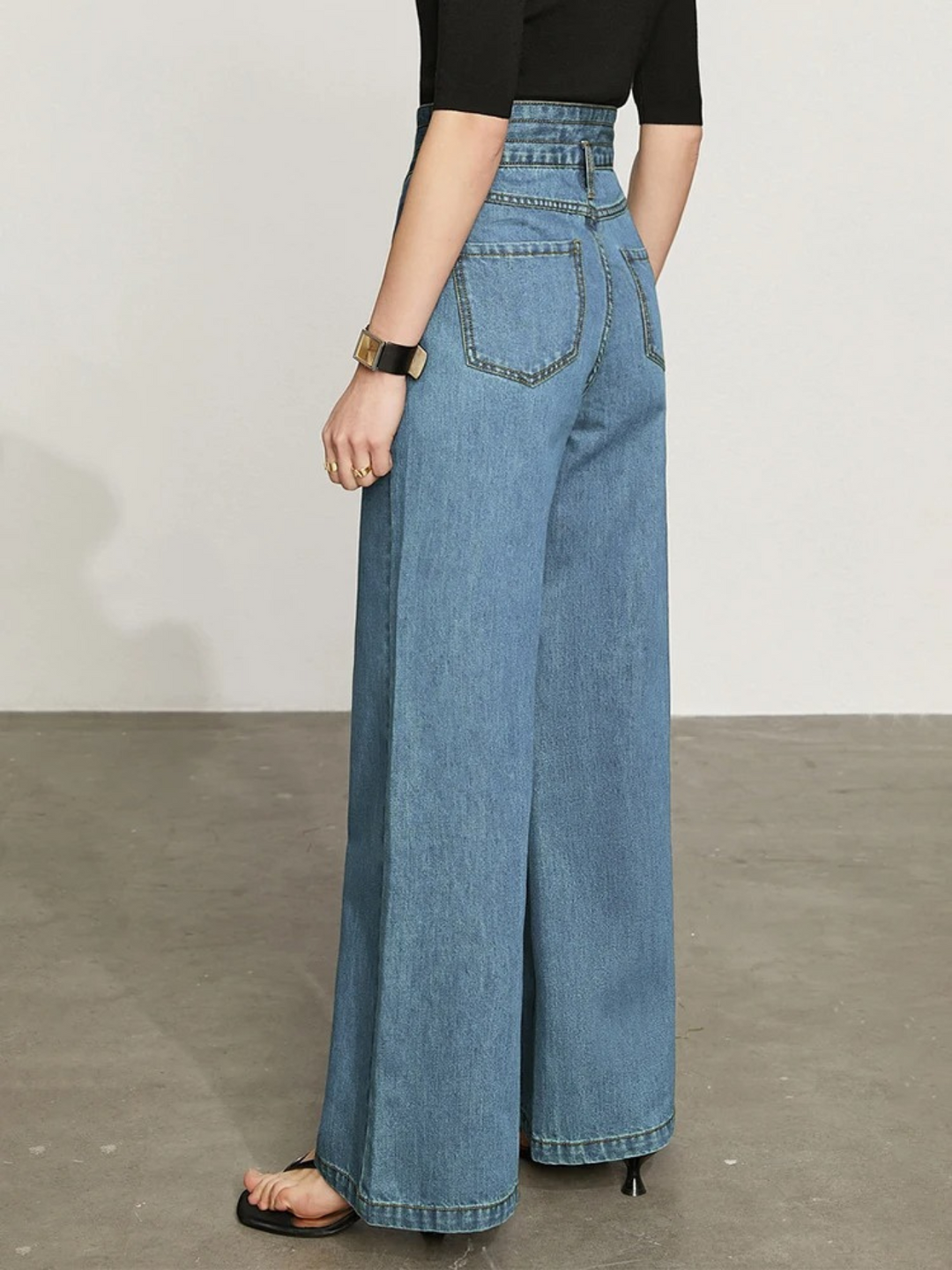 The back of  woman wearing a fitted black shirt with a pair of retro-inspired, wide leg jeans with a cinched waist design and a pair of sandals showcasing the pocket details of the jeans.