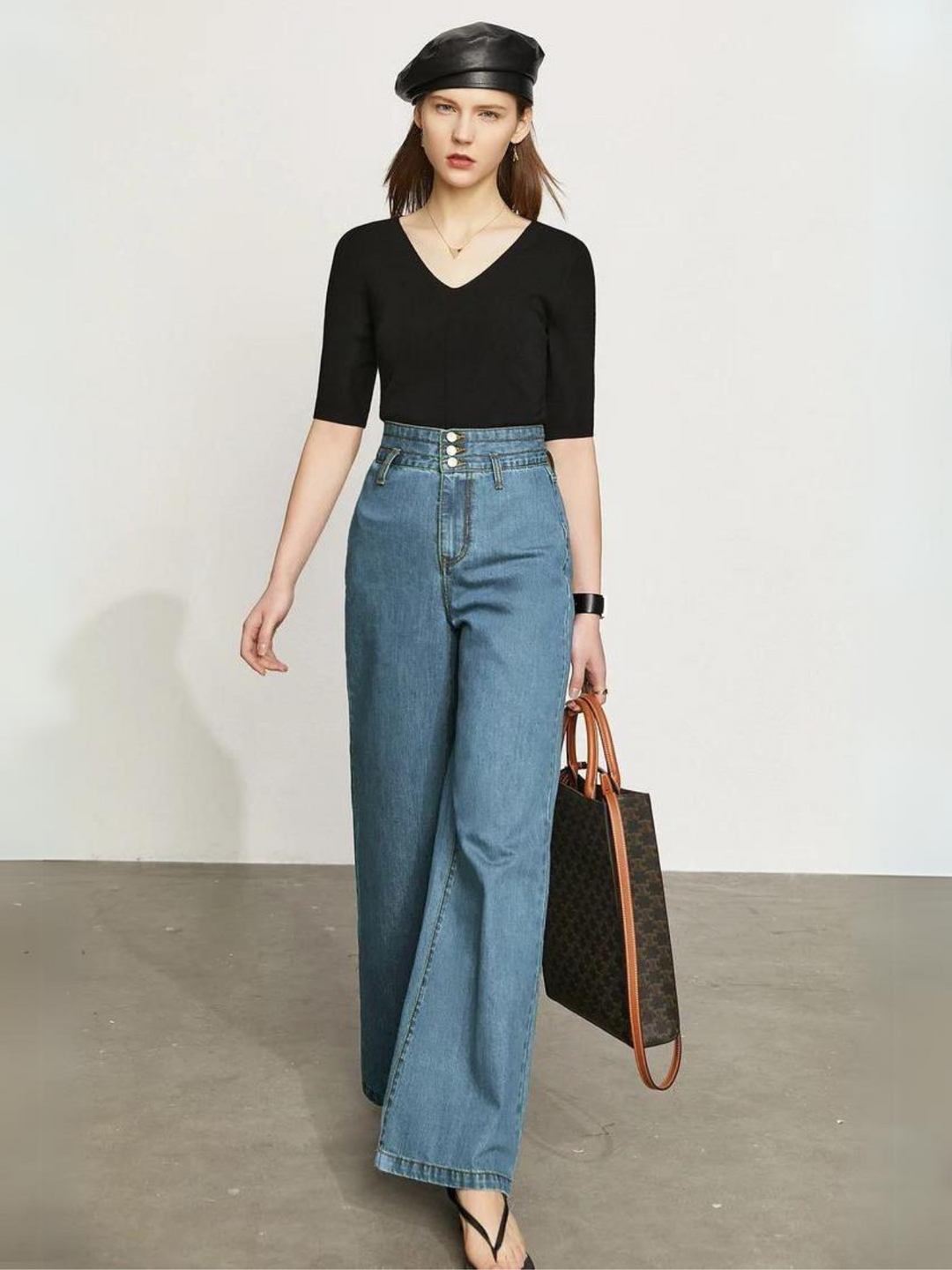 A stylish woman wearing a fitted black shirt with a pair of retro-inspired, wide leg jeans with a cinched waist design and a pair of sandals. She also has a black leather beret on for accessories.